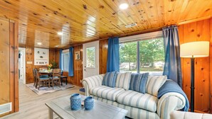 Ein gemütliches Zimmer mit Holzbalkendecke, einem gestreiften Sofa, einem Esstisch mit Stühlen und einem Fenster mit Vorhängen.