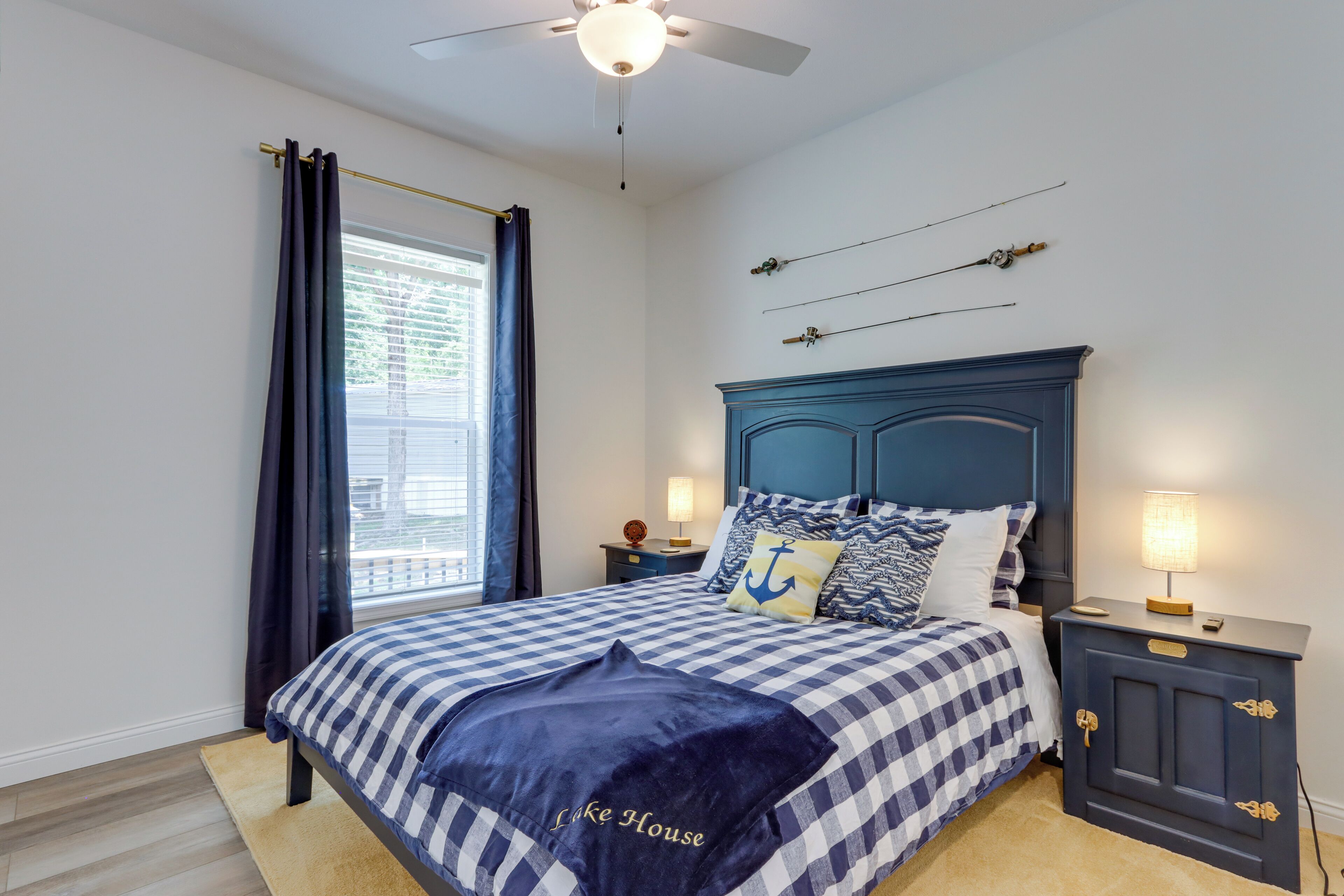 A bedroom with a checkered bedspread, a navy blue headboard, a nightstand with a lamp, and a window with curtains.