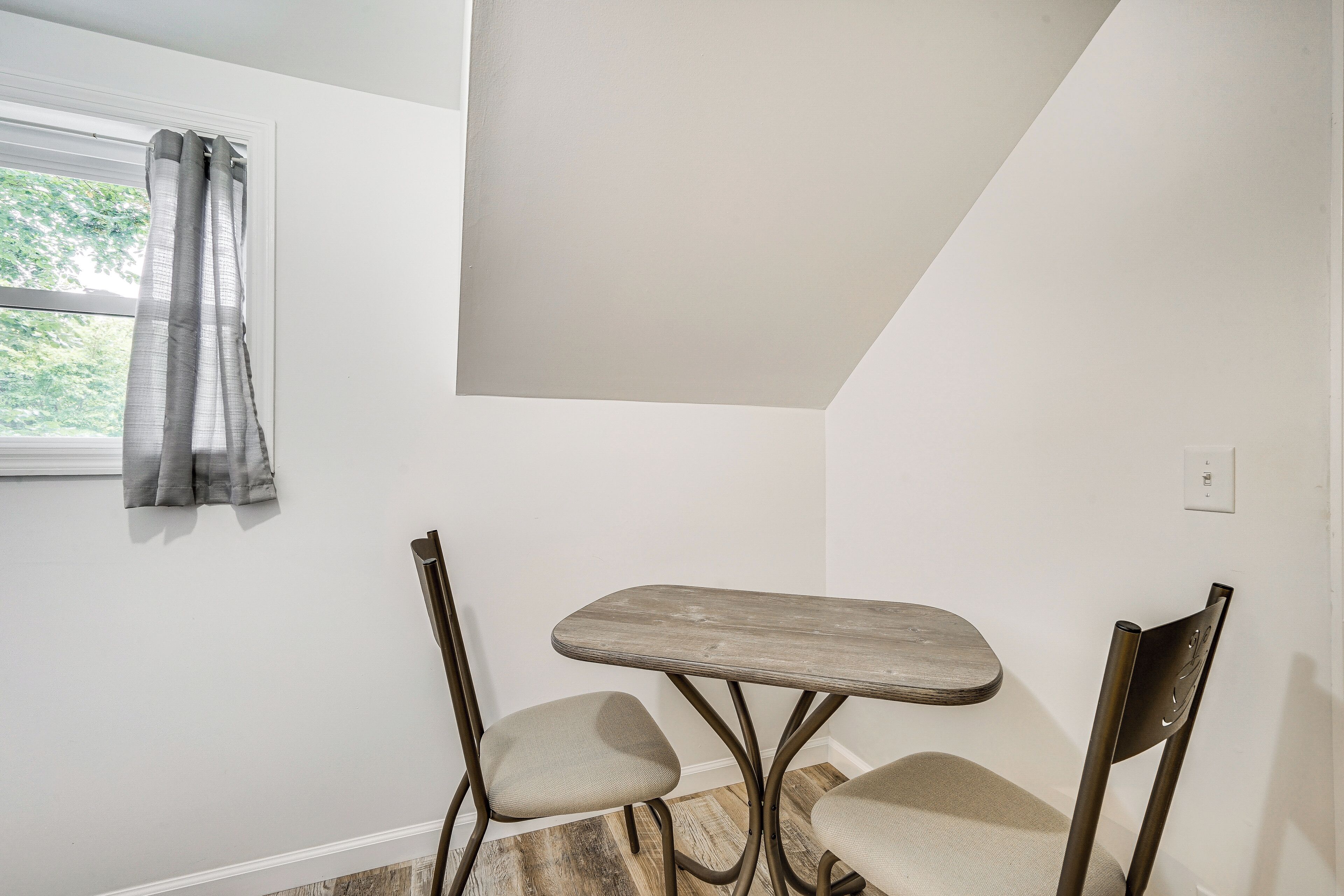 A small dining area with a table and two chairs, a window with curtains, and a white wall.