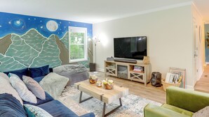 A living room with a large TV, a sofa, a coffee table with decorative items, and a mural of mountains and stars. - Black Mountain Retreat w/ Hot Tub + Mountain Views (Black Mountain)