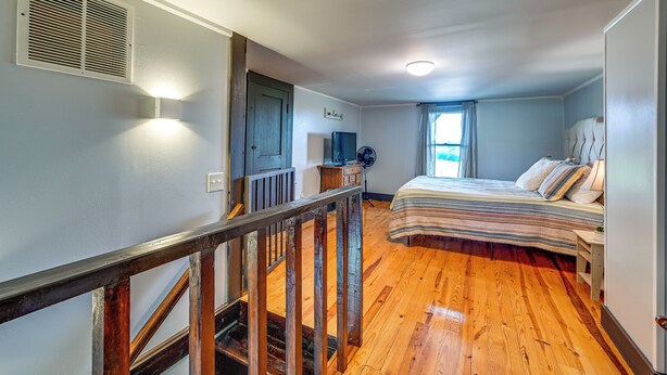 A bedroom with a wooden floor, a bed with striped bedding, a nightstand, a dresser, a window with curtains, and a staircase with a wooden railing. - Dobson Farmhouse w/ Scenic Porch - Near Vineyards! (Dobson)