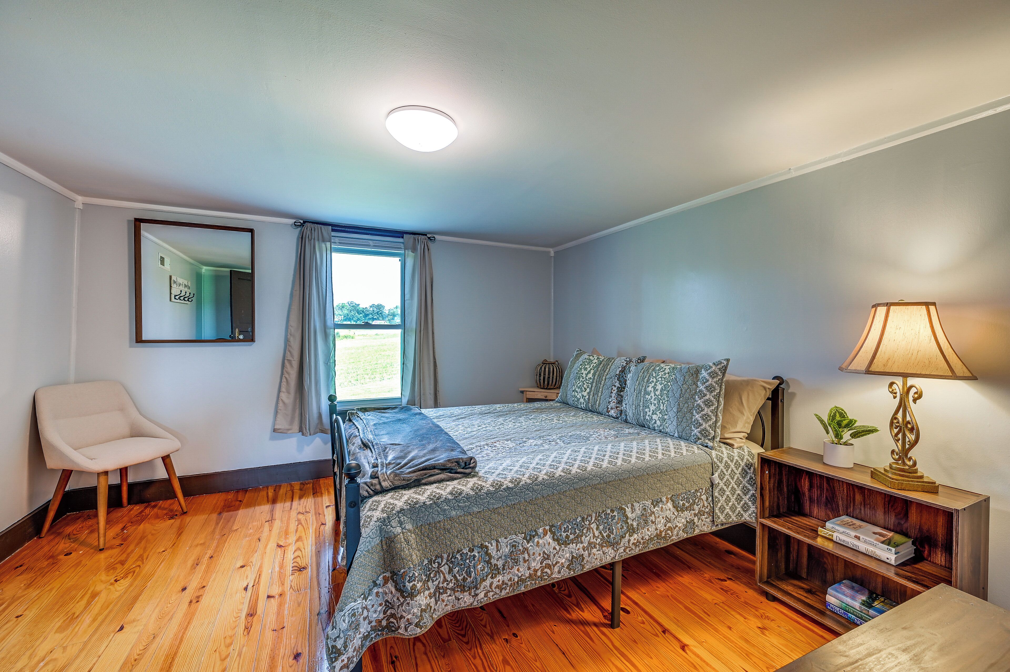 A bedroom with a bed, a chair, a bedside table with a lamp, and a window with curtains.