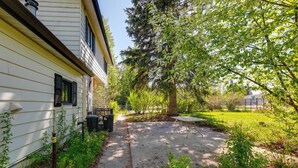 Un jardin à l’arrière d’une maison, avec une terrasse en béton, un arbre et une maison aux murs recouverts de bardage blanc et aux volets noirs.
