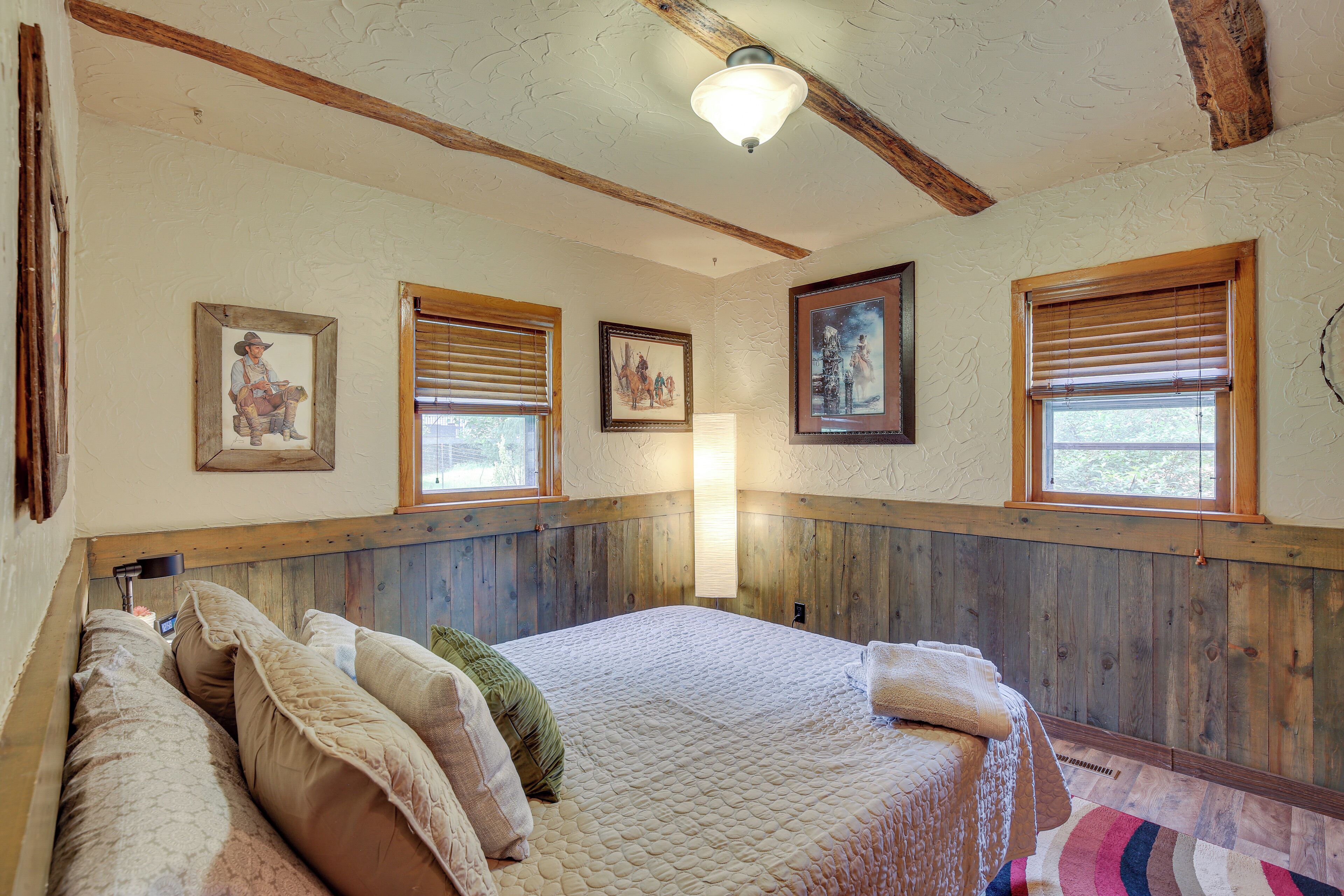 A bedroom with a large bed, wooden paneled walls, and three windows with blinds.