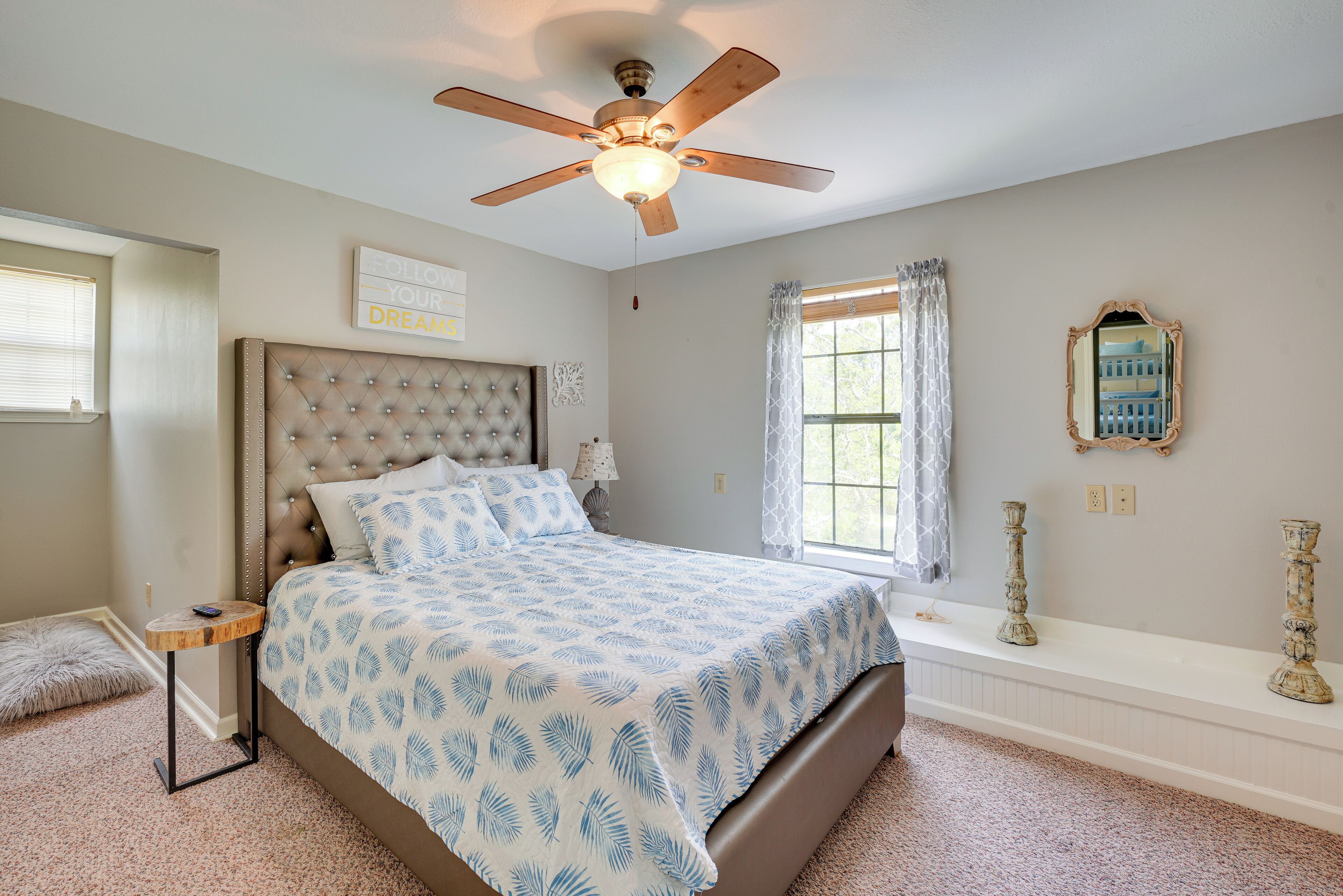 Une chambre à coucher avec une tête de lit capitonnée, un lit recouvert d’une couvre-lit à motifs de feuilles, un ventilateur de plafond, une fenêtre avec des rideaux, une table de chevet avec une lampe et un miroir sur le mur.