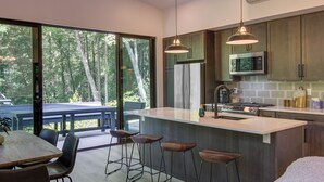 A modern kitchen with a large island, bar stools, and a view of the forest through large sliding doors. - Modern Cabin w/ Hot Tub & Fire Pit in Fairview! (Fairview)