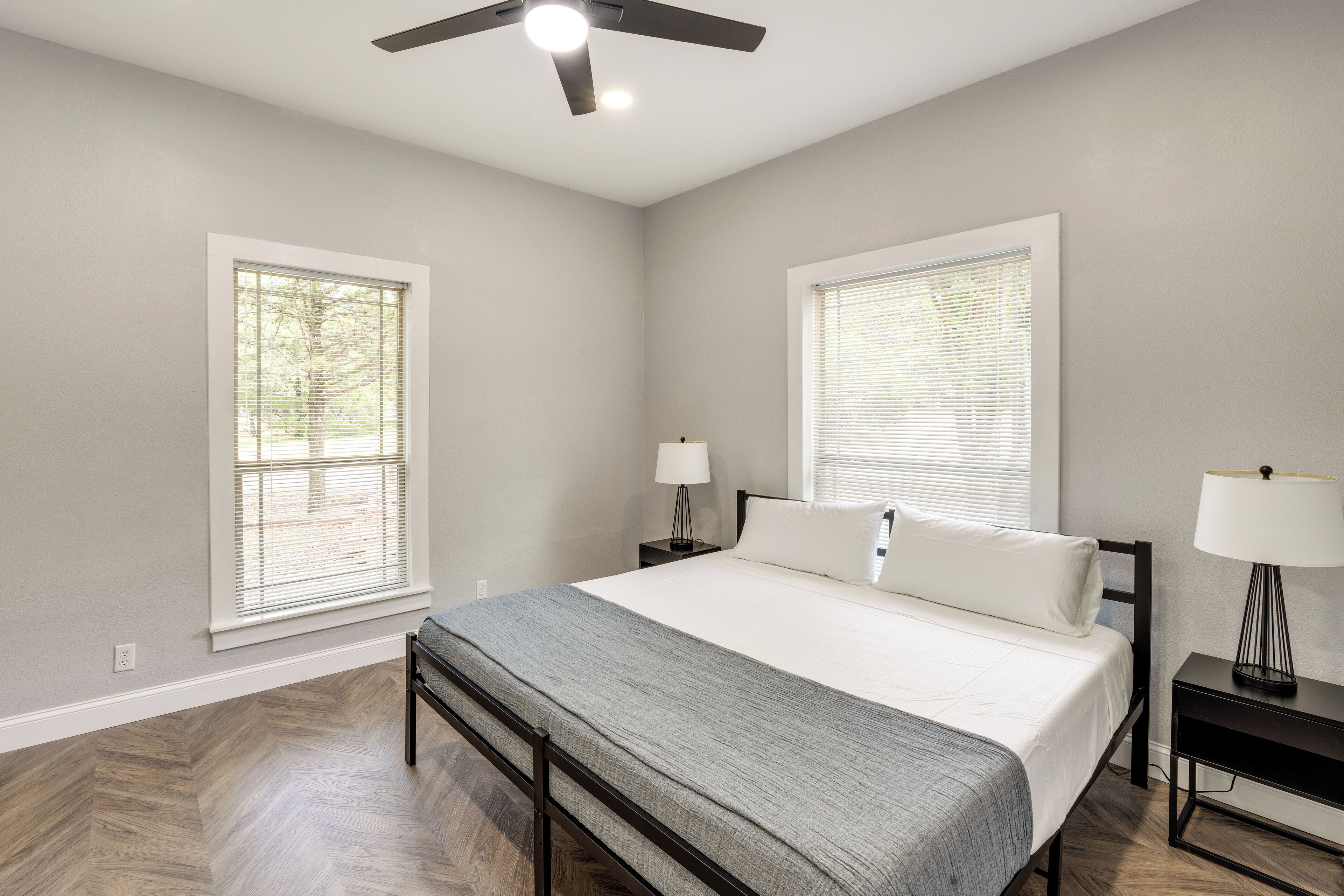 A bedroom with a bed, two nightstands, a ceiling fan, and large windows with blinds.