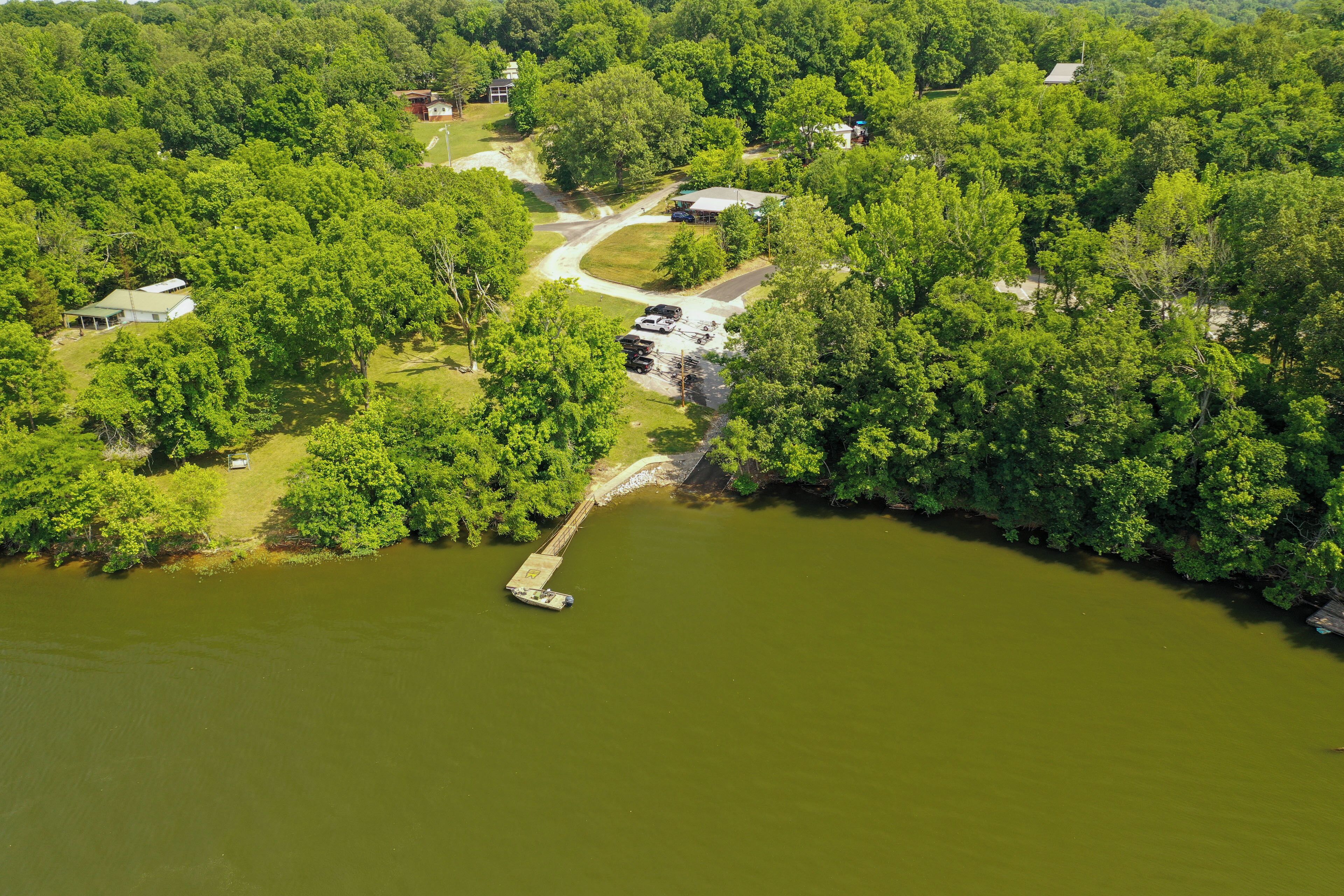 An aerial view of a lakeside property with a dock, surrounded by dense greenery and multiple buildings.