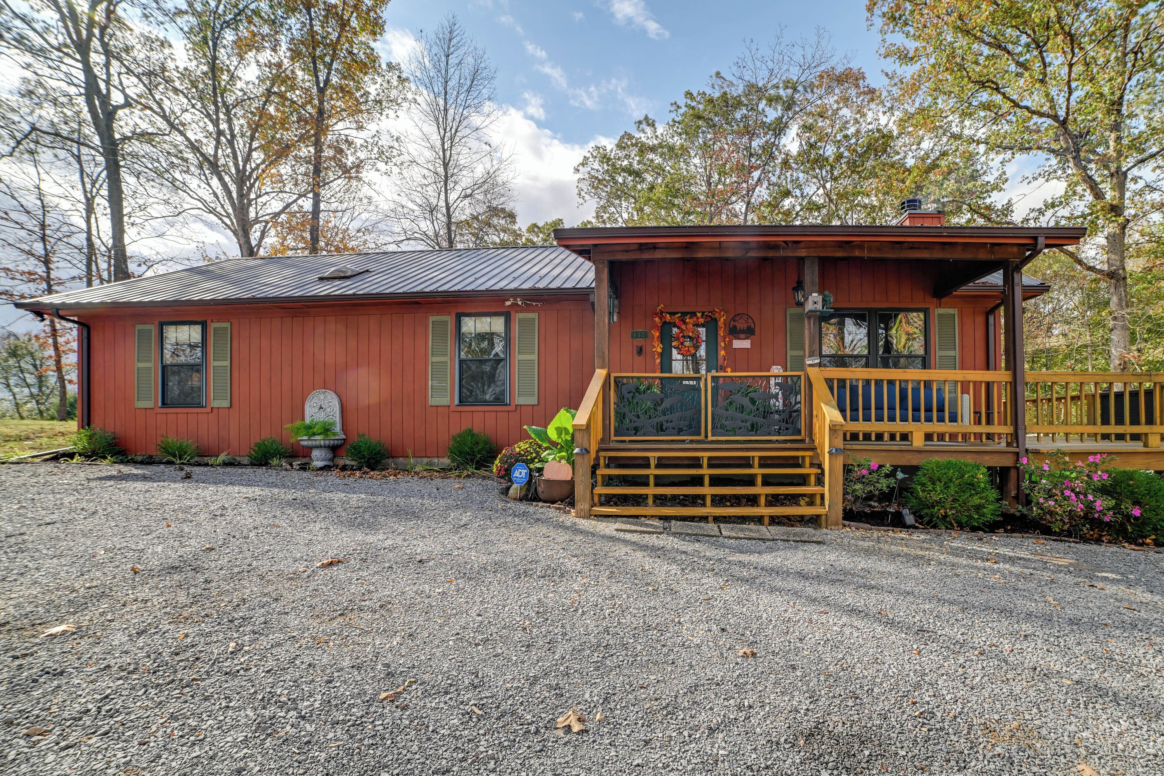 A small, single-story wooden cabin with a porch, surrounded by gravel and trees.