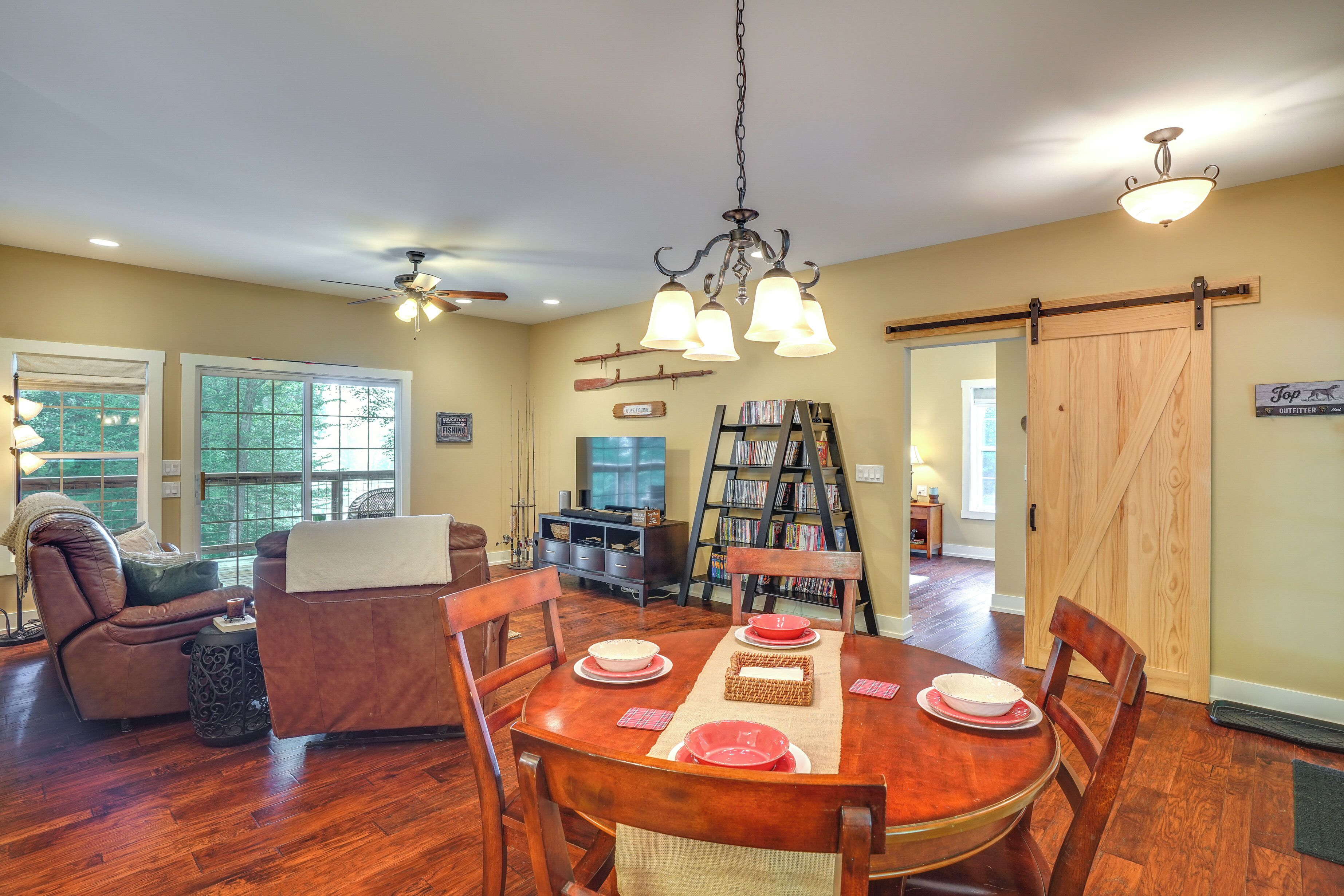 A dining area with a wooden table set for four, surrounded by chairs. There is a massage chair, a bookshelf, and a sliding barn door in the background.
