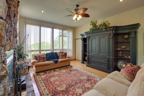A living room with a stone wall, a large window with blinds, a ceiling fan, a dark wooden cabinet, and a patterned rug. - Lake Ouachita Vacation Rental w/ Community Pool (Mount Ida)