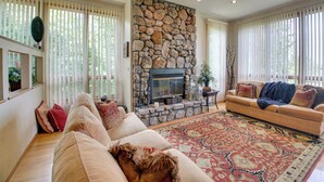 A spacious living room with a stone fireplace, a large window with blinds, a ceiling fan, and a patterned rug. - Lake Ouachita Vacation Rental w/ Community Pool (Mount Ida)