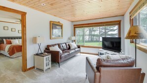 A spacious living room with a wooden ceiling, a large window with a view of trees, a leather sofa, a TV, and a bed with a headboard. - Minnesota Cabin w/ Deck & Private Beach (Merrifield)