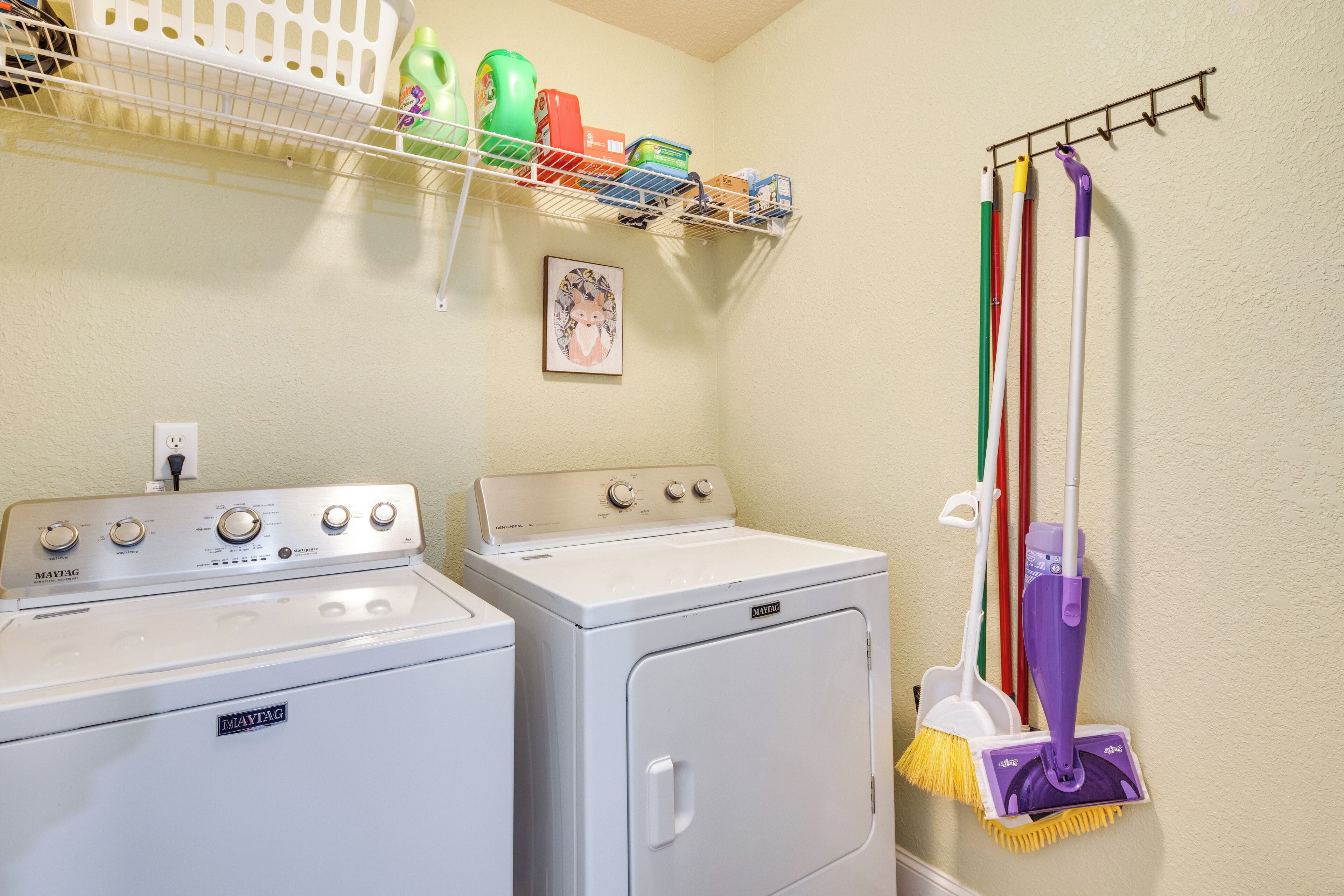 A laundry area with two white Maytag washers and dryers, a drying rack with colorful baskets, and a mop rack with various mops.