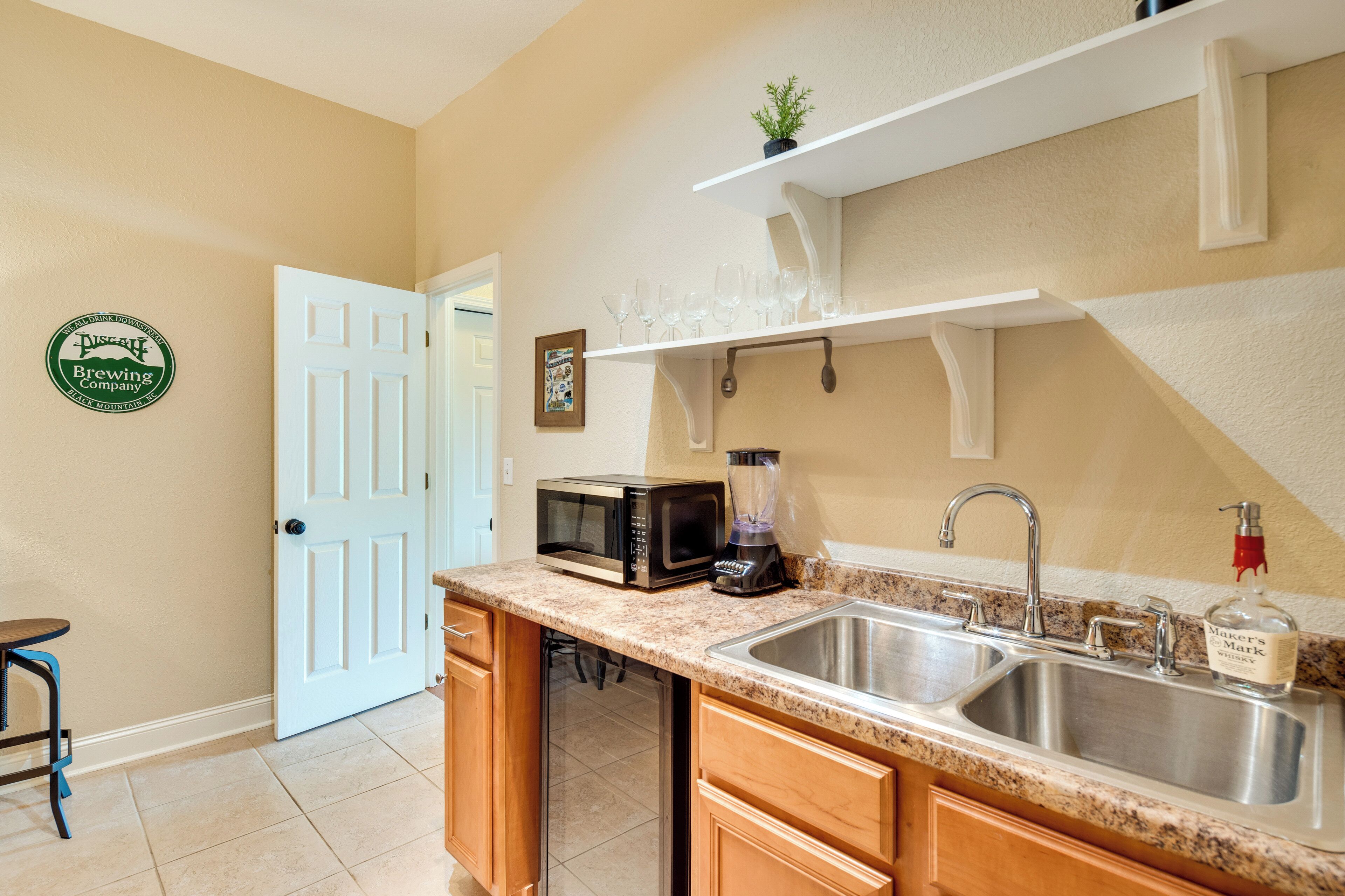 A kitchen with a double sink, a microwave, a blender, and a shelf with glasses.