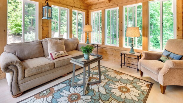 Una sala de estar acogedora con techo de madera, ventanas amplias, un sofá, un sillón y una mesita de noche con lámpara.