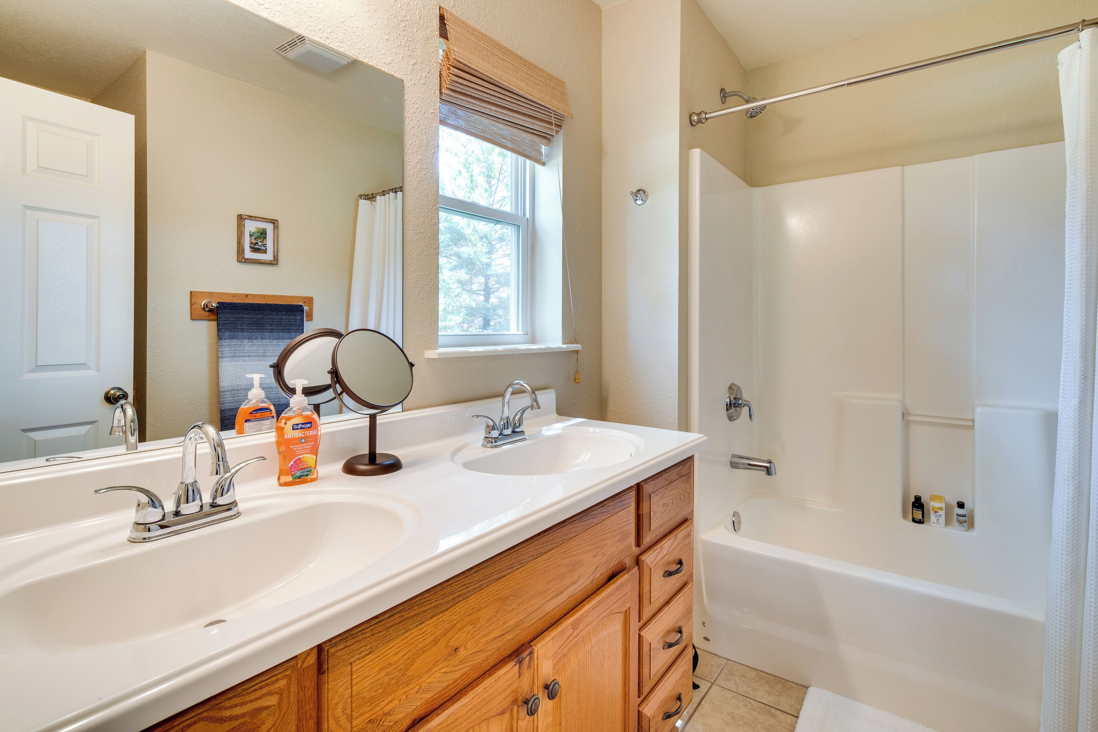 A bathroom with two sinks, a mirror, wooden cabinets, a window with blinds, and a bathtub.