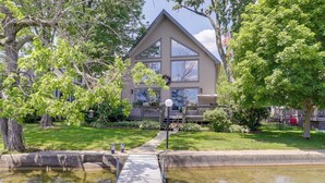 A two-story house with a large front yard, a wooden dock leading to a pond, and a well-maintained lawn. - Lakefront Vandalia Home w/ Boat Dock & Gas Grill! (Vandalia)