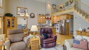 A spacious living room with a beige sofa, a leather recliner, and a white armchair. A wooden cabinet and a lamp are on a side table. A staircase leads to an upper level. - Lakefront Vandalia Home w/ Boat Dock & Gas Grill! (Vandalia)