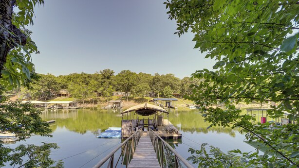 A wooden dock leading to a covered seating area by a calm lake, surrounded by trees and buildings. - Boat Dock & Fire Pit: Lakefront Paradise in Eucha! (Eucha)