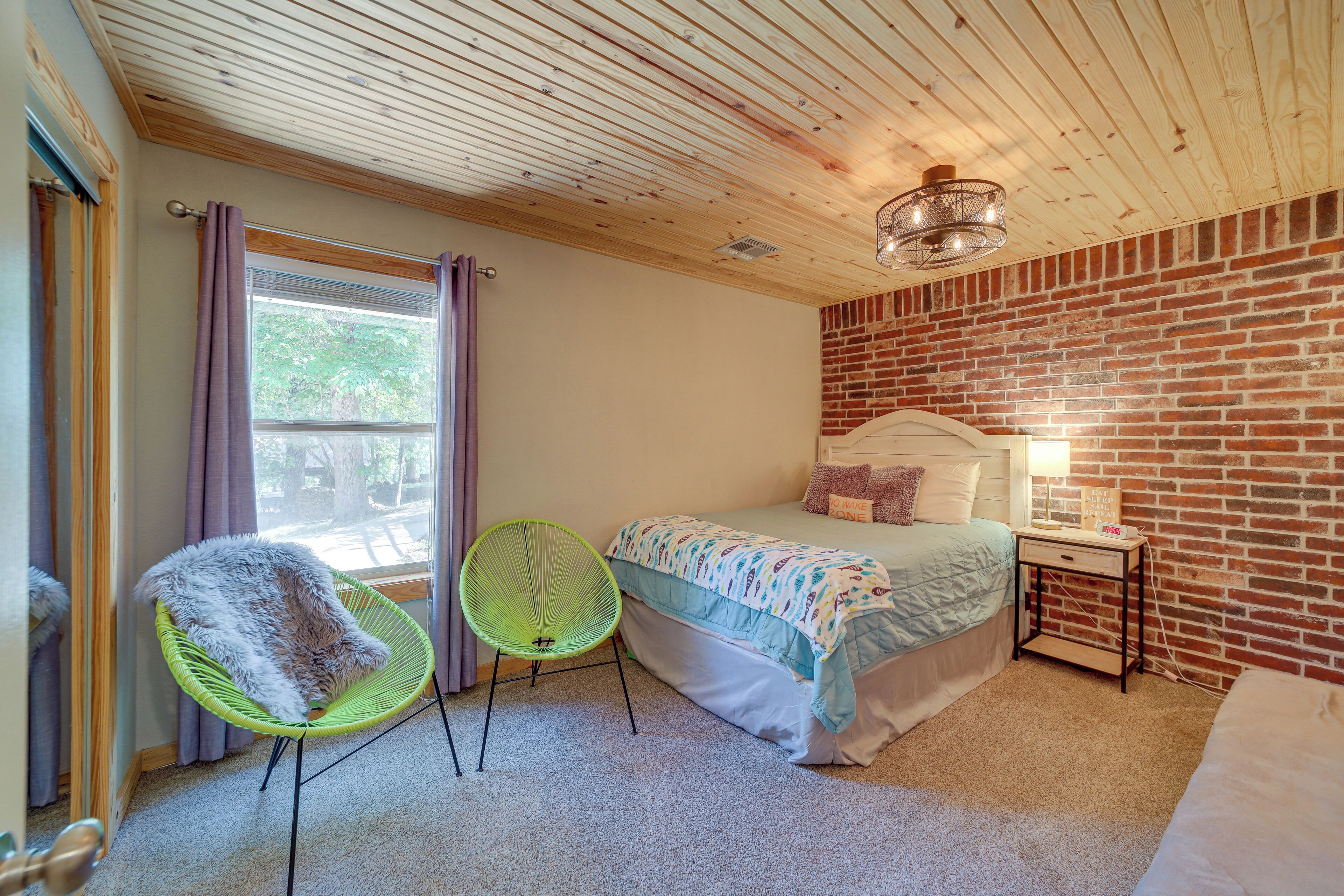 A bedroom with a brick wall, a bed with a white headboard, a green chair, and a small bedside table with a lamp.