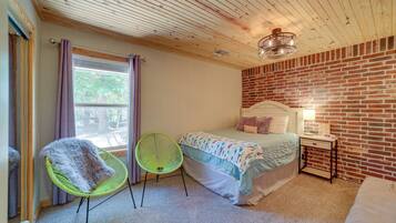 A bedroom with a brick wall, a bed with a white headboard, a green chair, and a small bedside table with a lamp.