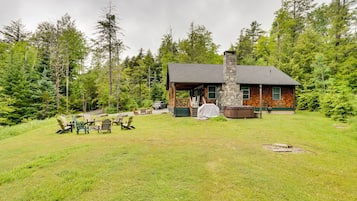 A cabin with a stone chimney, surrounded by a grassy yard with Adirondack chairs and a covered picnic table.