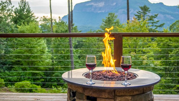 Ein Steinkamin mit zwei Weingläsern mit Rotwein auf einer Holzterrasse mit Blick auf eine gebirgige Landschaft.