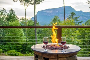 Uma lareira de pedra com dois copos de vinho tinto numa varanda de madeira, com vista para uma paisagem montanhosa.