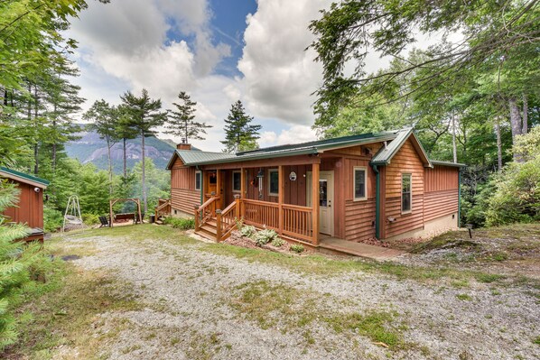 A rustic cabin with a porch, surrounded by trees and a mountain backdrop.