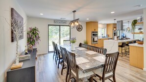 A modern kitchen with wooden cabinets, a central island, and a dining area with a glass table and chairs. - 6 Mi to Hunter Mtn: Winter Retreat w/ Hot Tub! (Tannersville)