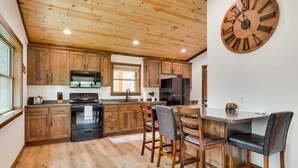 A kitchen with wooden cabinets, a large clock, and a bar area with stools. - Hatfield Vacation Rental w/ Private Hot Tub! (Merrillan)