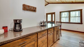 A kitchen with a granite countertop, wooden cabinets, a coffee maker, and a slow cooker. There is a window with a view of trees and a sign on the wall that reads "LAKE". - Hatfield Vacation Rental w/ Private Hot Tub! (Merrillan)