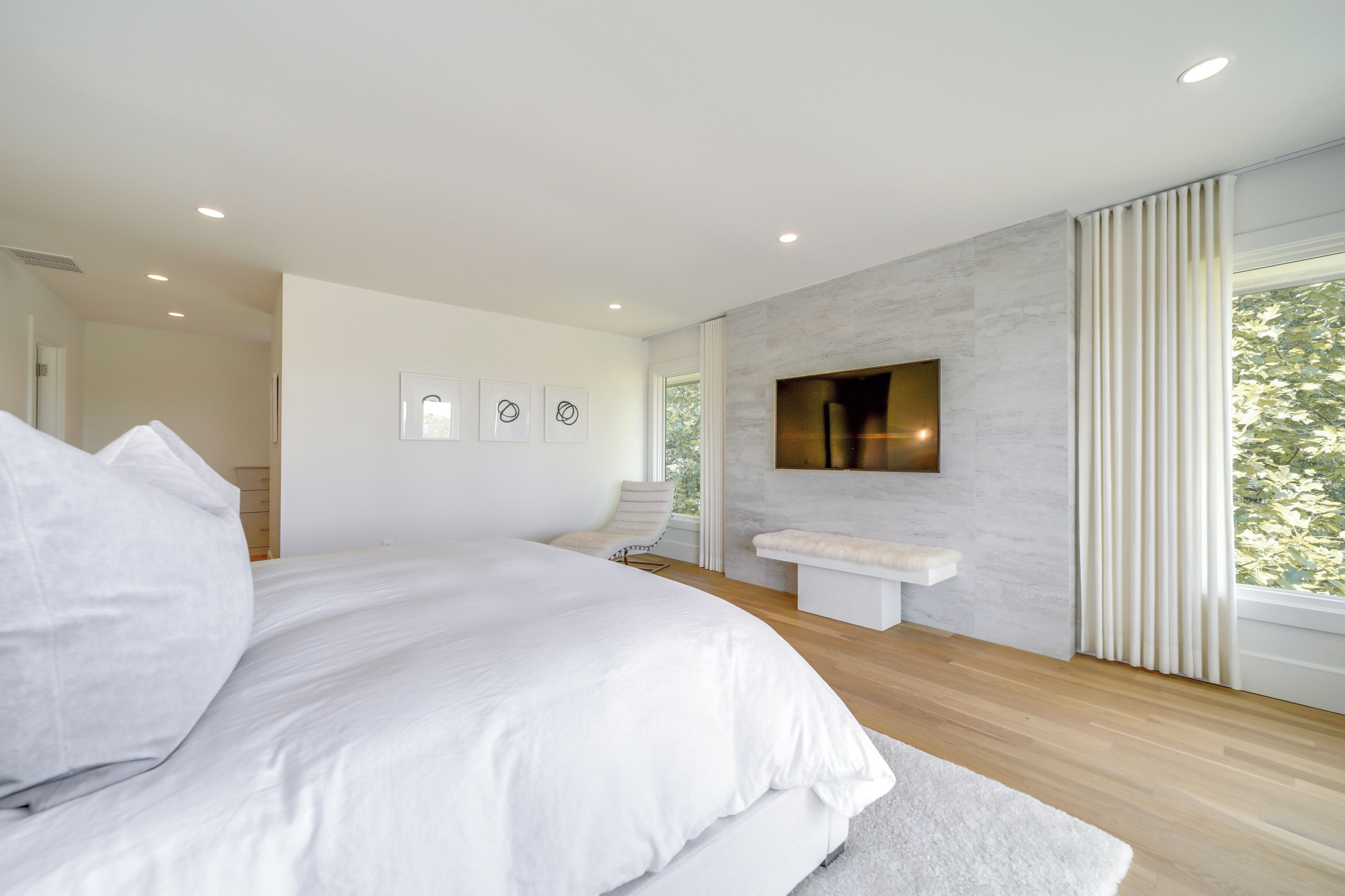 A modern bedroom with a large bed, a mounted TV, and a view of greenery through the window.