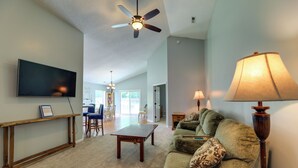 A living room with a ceiling fan, a TV mounted on the wall, a wooden coffee table, a green sofa with patterned cushions, and a lamp. - Swansboro Vacation Rental, 4 Mi to Hammocks Beach! (Swansboro)