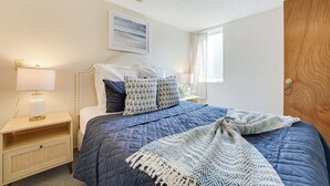 Una cama bien hecha con colcha azul, cabecera blanca y dos almohadas estampadas. Hay una lámpara de noche sobre una mesita de madera, y una ventana con cortinas a la derecha.