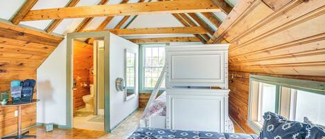 A bedroom with a wooden ceiling, a bed with patterned bedding, a white wardrobe, and a window with a view.