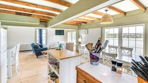 A spacious kitchen with wooden beams, a dining area with white furniture, and large windows offering a view of the outdoors. - Historic Winter Harbor Cottage w/ Waterfront Views (Winter Harbor)