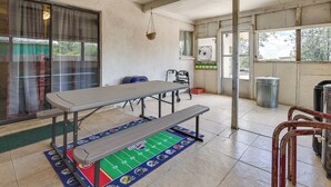 Um quarto com uma mesa grande e um banco, janela com cortinas, um ventilador e uma lixeira.