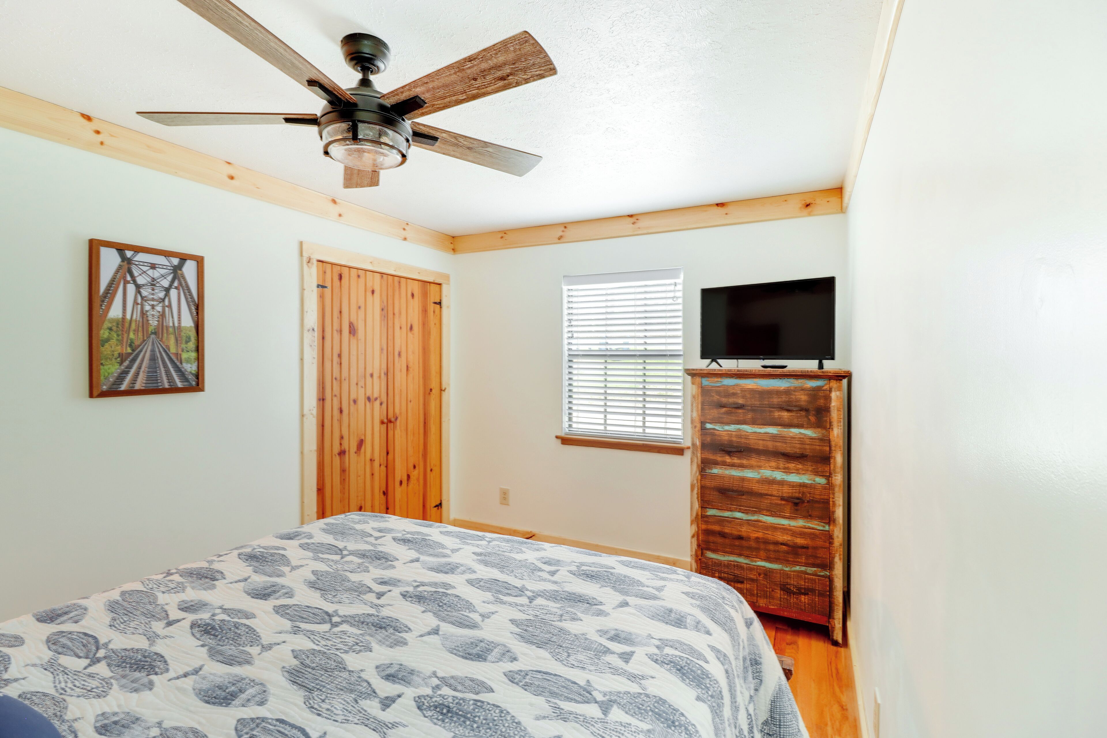 A bedroom with a bed, a ceiling fan, a wooden door, a window with blinds, and a television on a wooden dresser.