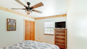 Um quarto com cama, ventoinha no teto, porta de madeira, janela com persianas e uma televisão sobre uma cômoda de madeira.