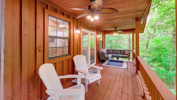 Um deck de madeira com duas cadeiras Adirondack brancas, um ventilador de teto e vista para uma floresta.