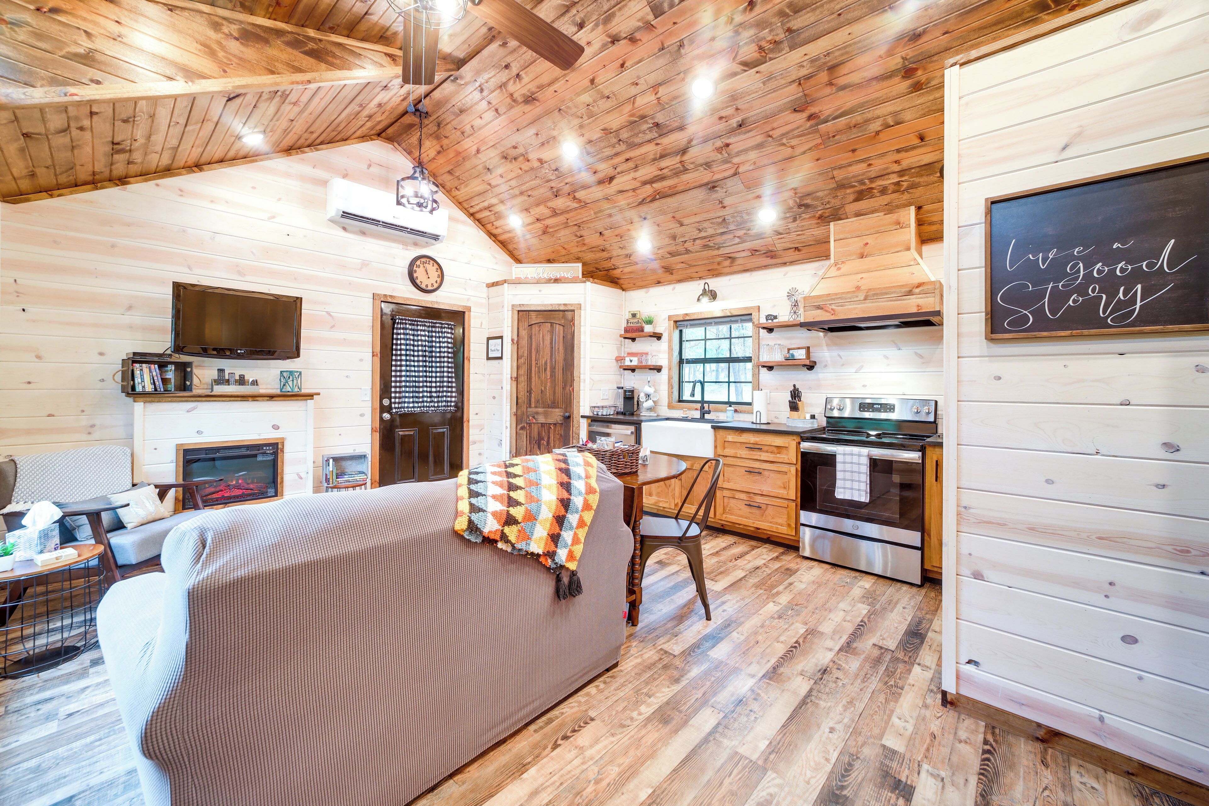 A cozy cabin interior with a wooden ceiling, a living area with a sofa and a TV, a kitchen with modern appliances, and a chalkboard with a motivational quote.