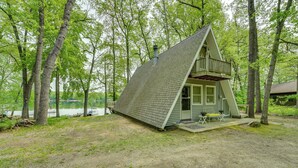 A small, A-frame cabin with a balcony, situated in a wooded area near a lake. - Long Lake Cabin w/ Kayaks, Private Beach & Dock! (Fountain)