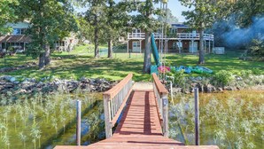 A wooden dock leading to a calm body of water with reeds, surrounded by trees and a building with a balcony in the background. - Lakefront Arkansas Escape w/ Grill, Dock & Kayaks! (Horseshoe Bend)