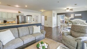 A spacious living room with a grey sofa, a matching armchair, and a coffee table with decorative items. A kitchen area with modern appliances and light green cabinets is visible in the background. - Lakefront Horseshoe Bend Home w/ Boat Dock! (Horseshoe Bend)
