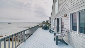 Une terrasse en bois avec une balustrade donnant sur une étendue d’eau, une table avec des chaises et un bâtiment doté de grandes fenêtres.