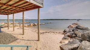 Une terrasse en bois, dotée d’une balustrade, surplombe une plage de sable parsemée de gros rochers et bordée par une étendue d’eau calme.