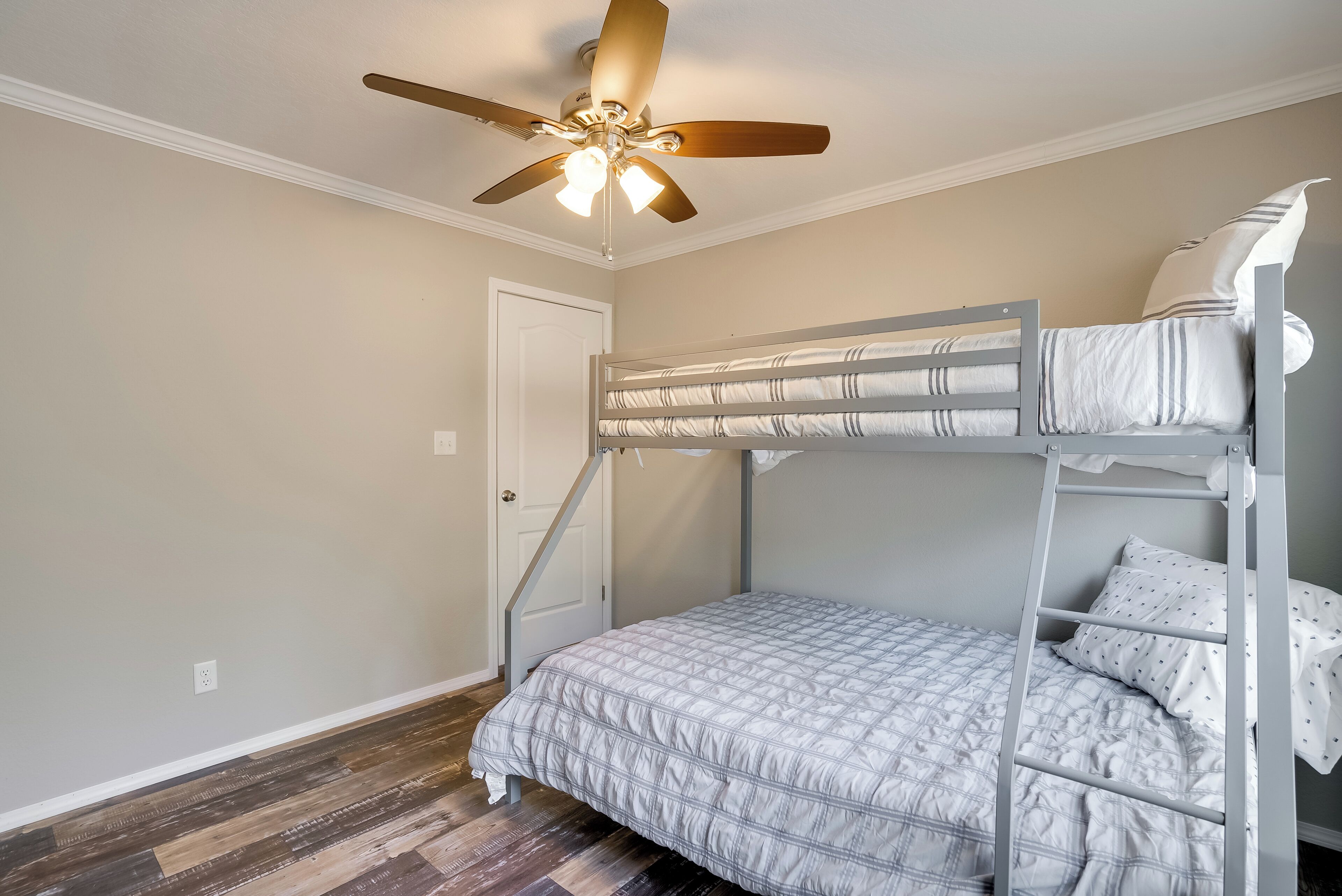 Quarto com beliche, ventilador de teto, piso de madeira e uma porta.