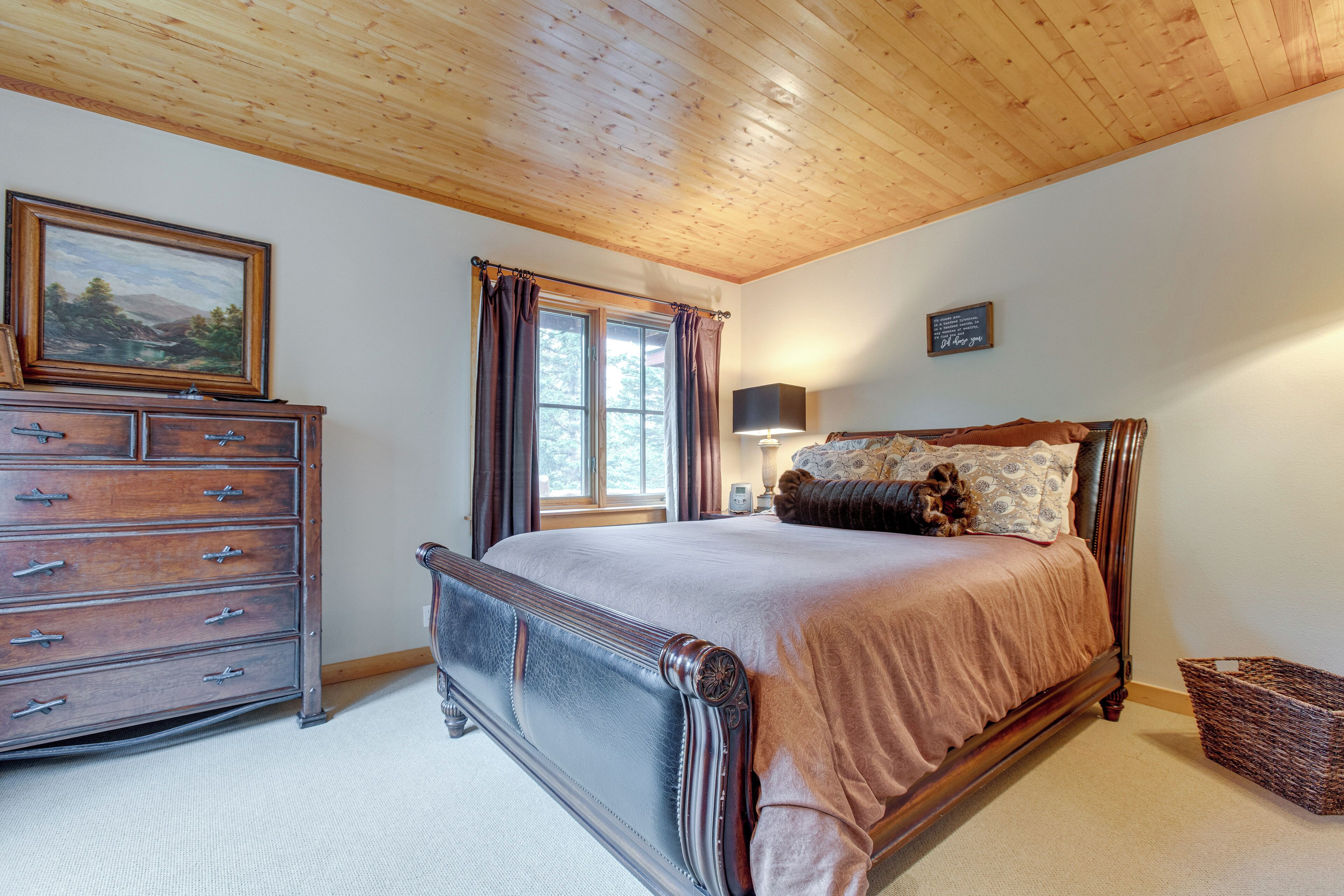 Een slaapkamer met een houten plafond, een bed met bruin beddengoed, een kast, een schilderij aan de muur en een raam met gordijnen.